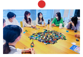 画像:学生たちが机を囲んで座り、台の上にあるたくさんのレゴブロックを、学生たちが手に取りながら話している様子